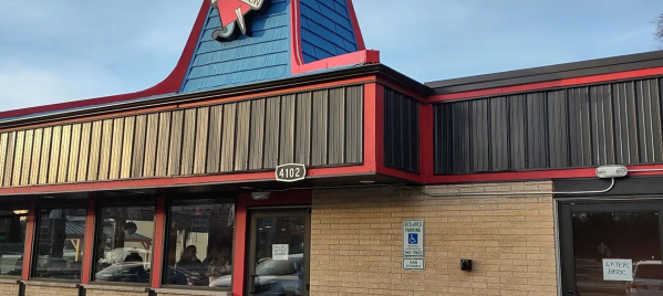 Exterior of a retro-style diner with beige brick walls, red and black trim, and a blue gabled roof. A large sign featuring a heart and a woman in a red dress sits atop the building. A handicapped parking sign is visible near the entrance.
