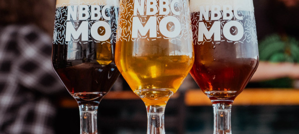 Three full beer glasses, filled to the brim with foam at the top of each one, on a wooden table tap and people eating and drinking in the background.
