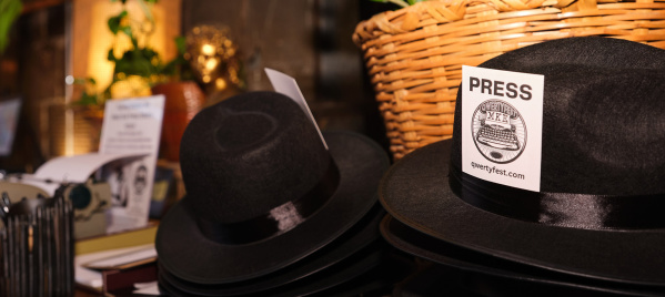 Stacks of black fedora hats labeled ‘PRESS’ with QWERTYFEST MKE tags on a wooden table decorated with a vintage typewriter, papers, and a wicker basket with green plants in the background.