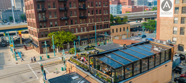 The Lux Skyhaus at Cafe Benelux, a glass greenhouse rooftop dining space in Milwaukee, overlooks a pedestrian plaza with teal lamp posts. The transparent structure reveals lush greenery and diners inside, surrounded by historic red brick buildings and downtown Milwaukee's skyline.