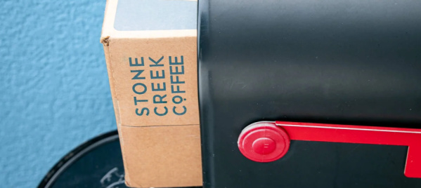 Cardboard box labeled "Stone Creek Coffee" placed inside a black mailbox with a red flag, against a blue textured wall.
