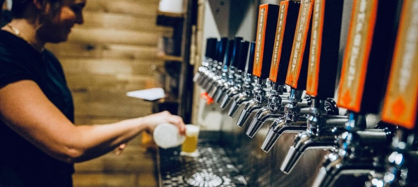 Person pouring a draft beer from a row of shiny metal taps at a bar with wooden walls and warm lighting.