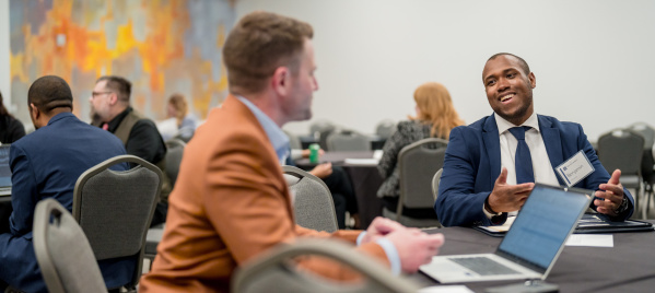 Two business professionals conversing at a conference table - one in an orange blazer with back to camera, the other in a navy suit smiling and gesturing while speaking. Multiple attendees visible in background conference room setting.