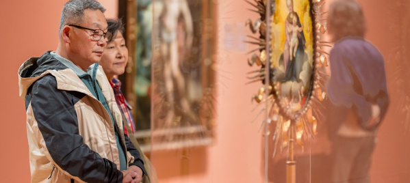 A middle-aged man wearing glasses and a tan-and-black jacket stands with his hands clasped, looking thoughtfully at an artwork in what appears to be an art museum or gallery. Behind him, a woman stands slightly out of focus, also observing the exhibit.

In the foreground on the right, there is a blurred religious-style artwork or icon displayed in a glass case, with a gold decorative frame. The overall scene has warm lighting and a calm, contemplative mood.