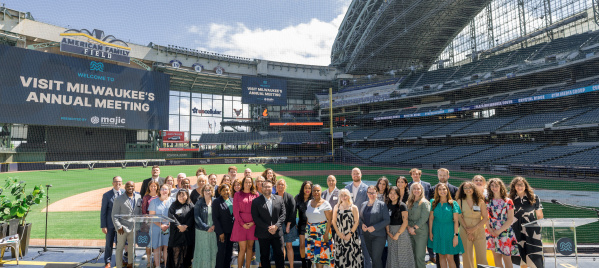 Group photo of attendees at Visit Milwaukee's Annual Meeting, posed on a stage at American Family Field (Milwaukee Brewers stadium) with stadium seating and banners visible in the background.