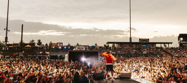 A performer in a red hoodie stands on an outdoor stage facing a massive crowd at sunset. Thousands of fans pack the venue, many holding up phones to capture the moment, while bright stadium lights and food stand signs glow in the background.