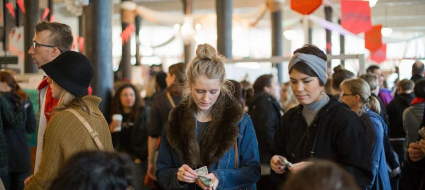 A busy indoor market scene with people browsing and making purchases. Two women in the foreground count money, while crowds of shoppers fill the background among vendor booths and hanging signs.