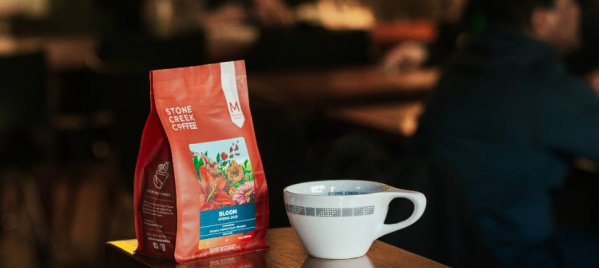 Bag of Stone Creek Coffee and a white ceramic coffee cup on a wooden counter, with a blurred café setting in the background.