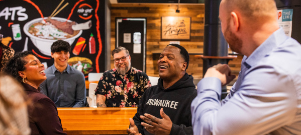 LeRoy Butler is wearing a black Milwaukee hoodie speaks animatedly while seated at a table inside 3rd Street Market Hall, with several people listening and smiling around him
