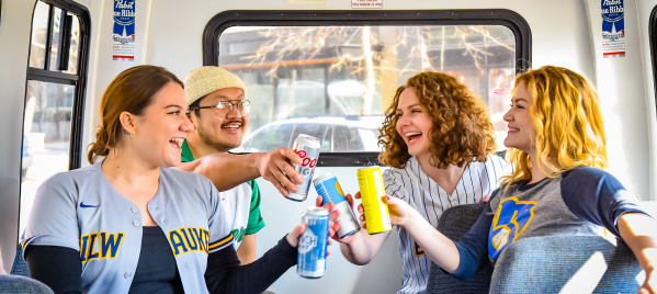 Four friends sitting inside a shuttle bus, smiling and clinking their drinks together. Two are wearing Milwaukee sports jerseys, creating a festive and celebratory atmosphere.