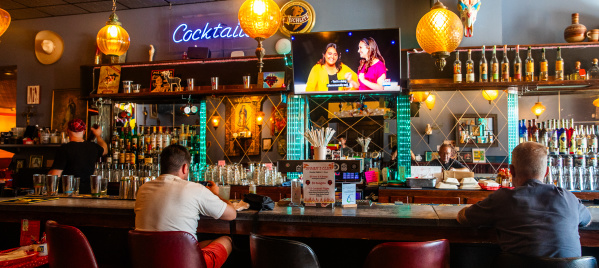 People are seated at a colorful, retro-style bar with eclectic decor, warm lighting, and a neon "Cocktails" sign glowing above shelves stocked with liquor bottles. A television above the bar shows a lively talk show.