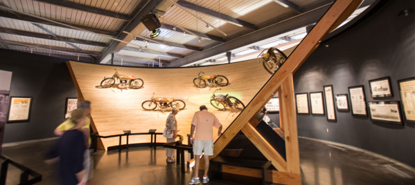 Visitors explore a motorcycle exhibit inside the Harley-Davidson Museum. The display features vintage motorcycles mounted on a large wooden wall with a dramatic angled support beam, and framed historical documents and images line the surrounding walls.