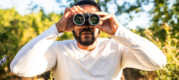 A man in a white long-sleeved shirt holds binoculars up to his eyes while standing outdoors in a sunny, tree-filled setting with green foliage and dappled sunlight in the background.