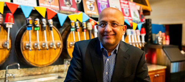 Sharad Chadha, CEO of Sprecher Brewing Company, sits smiling in a dark suit and blue checkered shirt at the brewery's taproom bar. Behind him are multiple beer taps, colorful triangular bunting, and shelves displaying various Sprecher product bottles and signage.