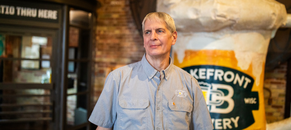 Russ Klisch wears a light blue button-down shirt at Lakefront Brewery. Behind him are exposed brick walls, industrial-style lighting, and a large format frothing beer mug.