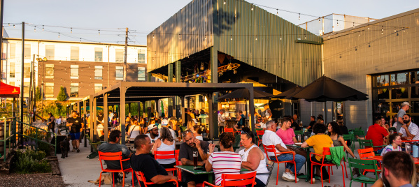 A large patio at a brewery is full of people sitting in red chairs. The golden light of sunset spills over the space. A large bar is in the background with a green overhang.