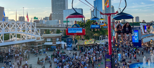 Crowds enjoying a lively outdoor festival with food stands and a sky ride, with people seated on chairlifts overlooking the scene and city skyline.