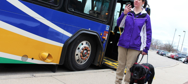 A woman with a visual impairment exits a city bus while holding a harness attached to her black guide dog. She is wearing a purple jacket and beige pants, and is using a smartphone as they walk along the sidewalk.