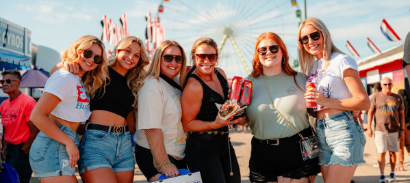 Six women wearing sunglasses pose together with drinks, fair food and a box of Original Cream Puffs in front of the Ferris wheel at the Wisconsin State Fair.