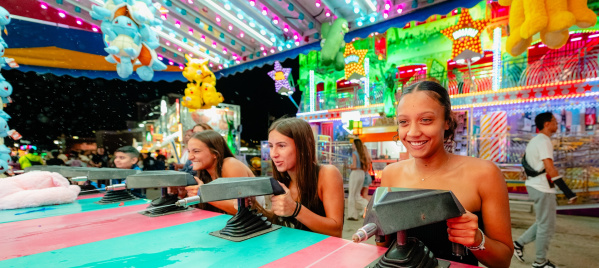 Three women smile while competing at a carnival squirt gun game beneath colorful lights and oversized stuffed animal prizes at the Wisconsin State Fair.