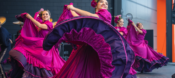 A group of female dancers performs a traditional Mexican folklórico dance on stage. They wear vibrant magenta dresses with purple ruffles and floral headpieces. The lead dancer in the foreground smiles brightly while gracefully twirling her flowing skirt. The dancers behind her mirror the movement, creating a colorful, energetic scene against a modern stage backdrop.