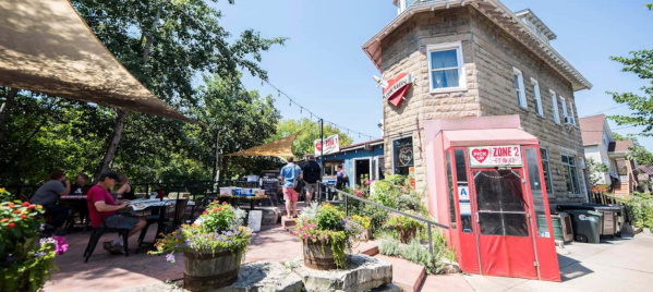Outdoor patio scene at a charming café with a stone tower-style building, red phone booth, and colorful flower planters. Diners sit at tables under shade sails, surrounded by lush greenery on a sunny day.