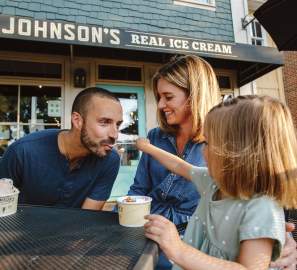 A young daughter feeding ice cream to her dad while her mom watches on the front patio of  Johnson's Ice Cream