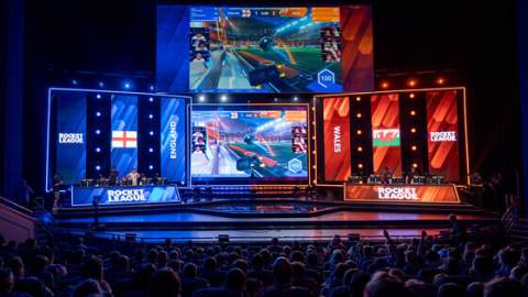 A crowd sit in darkness in a large venue in front of a stage lit in bright blue and red, ready to host an esports event