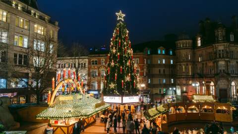 A tall Christmas tree stands in a square in Birmingham at night, the lights are lit and people are gathered round its base at Christmas market stalls