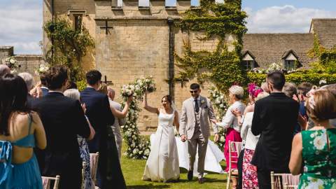 A couple smile at guests as they walk down the aisle at their outdoor wedding ceremony in the gardens of Ellenborough Park