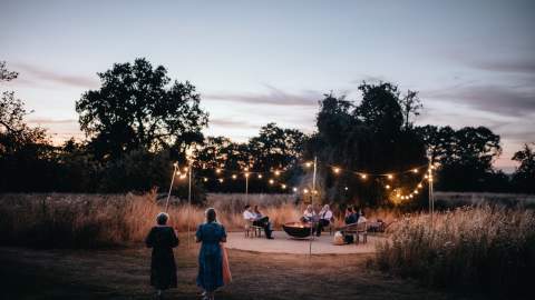 As the sun sets, a group of wedding guests relax around a fire pit in wicker chairs, surrounded by festoon lights at Elmore Court