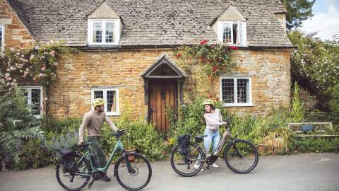 A couple push their bikes past a beautiful old cottage in a Cotswold village