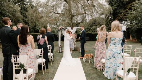 A bride and groom exchange vows at an outdoor ceremony, watched by their wedding guests and a Cavalier King Charles spaniel