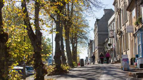 Dappled sunlight casts shadows on the pavement as people walk along High Street, Witney in the Cotswolds