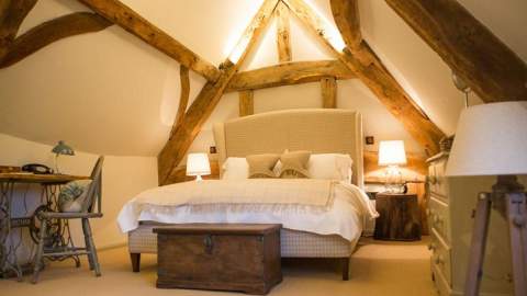 An upholstered large bed sits beneath original wooden beams on the ceiling and walls at The Porch House in the Cotswolds
