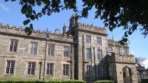Historic stone building with crenellated roofline and tall windows, labeled "The Bedford." Framed by tree branches, evokes a sense of timeless elegance.