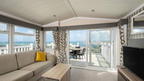 Bright living room with a beige sofa and yellow cushion, facing sliding glass doors that open to a balcony with a sea view. Airy and serene atmosphere.