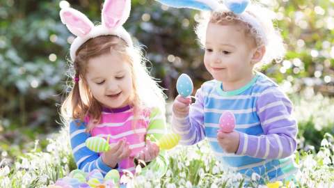 Two young children in striped shirts, wearing bunny ear headbands, joyfully play with colourful Easter eggs in a sunny, flower-filled garden.