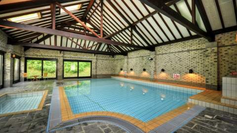 Indoor pool with rustic wooden beams and brick walls, sunlit through large windows. The pool is calm, with seating nearby, creating a serene atmosphere.