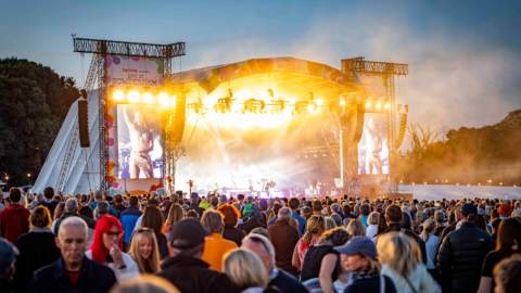 A large crowd of people enjoying an outdoor concert, with colourful lights and a stage in the background.