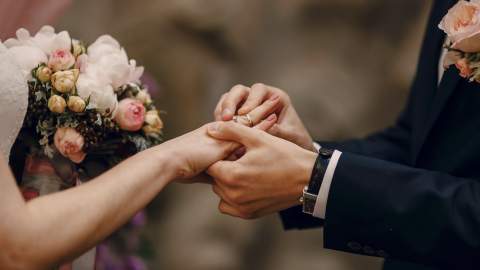 A groom places a ring on the bride's finger during their wedding ceremony. The bride holds a bouquet of pink and white flowers.