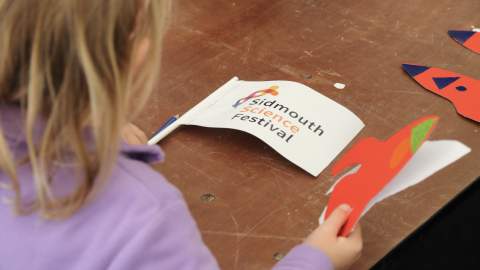 A child in a purple sweater holds a colorful paper rocket and a flag with "Sidmouth Science Festival" on a wooden table, conveying a playful, educational atmosphere.