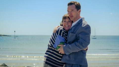 A couple embraces warmly by the beach under a bright blue sky. Behind them, the calm sea and a distant sailboat set a peaceful, joyful tone.