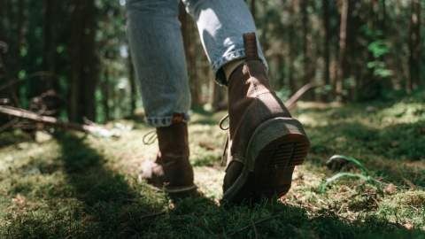 Close-up of a person wearing brown boots and blue jeans walking on a sunlit, moss-covered forest floor, conveying a sense of adventure and tranquility.
