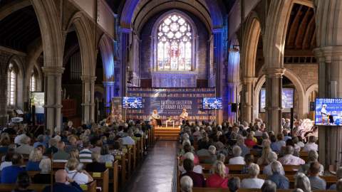 A large audience fills a historic church with tall arches and stained glass windows, attending a literary festival. The atmosphere is focused and attentive.