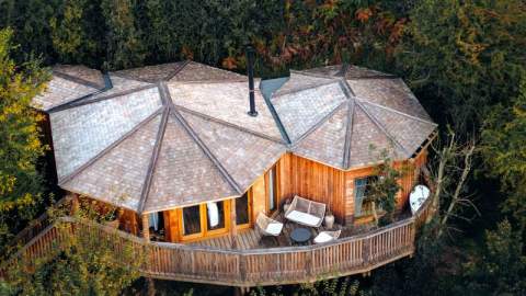 Aerial view of a wooden treehouse with hexagonal roofs, nestled among lush greenery. A cosy deck with chairs suggests relaxation and tranquility.