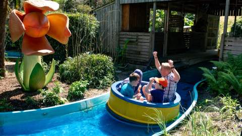 A group of people enjoy a log flume ride, passing by a large, whimsical orange flower sculpture. Lush greenery surrounds the area.