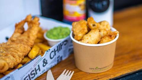 A hearty serving of fish and chips with mushy peas in a newspaper-lined box on a wooden table. A paper cup with fried snacks is nearby, evoking a warm, casual dining experience.