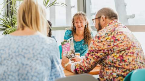 A group of people seated around a table in a bright, modern indoor setting with large windows and natural light. One person is holding a colorful brochure titled “Old Town Guided Tour” with images and text visible on the cover. The table has an orange surface, and there are green plants in the background.