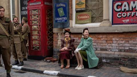 Actors dressed in 1940s clothing sit beside a vintage British red telephone box on a cobbled street, surrounded by period military uniforms and retro advertising signs.